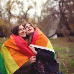 фото однополой женской пары, девушки обернулись в прайд-флаг и смотрят вверх