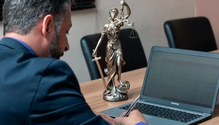 мужчина (прокурор) записывает что-то ручкой, сидя перед открытым ноутбуком, на столе рядом также стоит статуя богини правосудия Фемиды