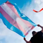 человек несёт транс флаг на фоне неба, поверх изображения красная лента - символ ВИЧ