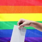 Рука опускает лист в урну на фоне радужного флага. ЛГБТ, голосование, выборы