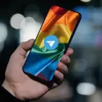 Рука держит телефон, на котором изображён прайд-флаг и поверх него иконка Телеграма