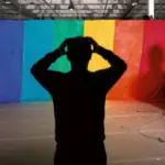 тёмная фигура в ужасе хватается за голову перед ярким восьмицветным радужным полотном