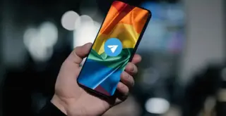 Рука держит телефон, на котором изображён прайд-флаг и поверх него иконка Телеграма